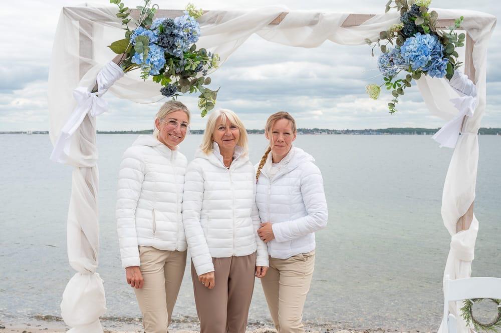 Gaby Löwel mit Team am Strand vor einem dekorierten Traubogen bei einer romantischen Strandtrauung in Strande